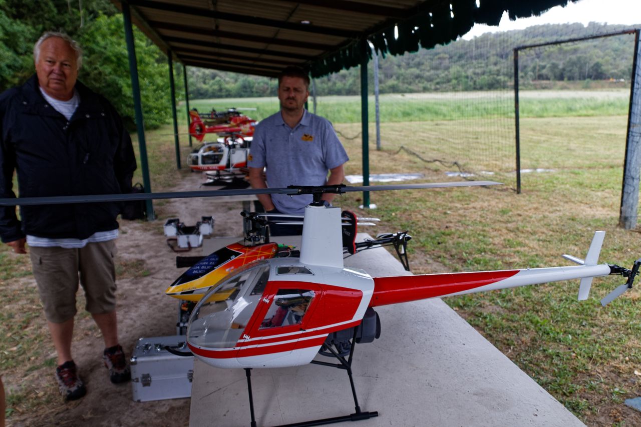 Two men showcasing remote control model helicopters under a pavilion in a field.